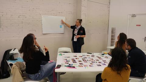 L'animatrice du serious game, debout, pointe du doigt une feuille blanche accrochée sur un mur, tandis que des étudiantes, étudiants et professionnels l'écoutent, assis autour d'une table sur laquelle ils ont crée une frise. 